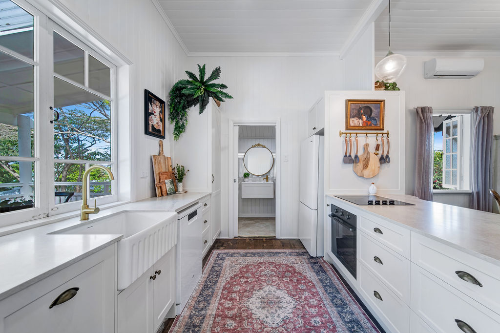 The kitchen after renovation, with white cabinetry, brass tapware, restored timber floors and a view through to the vanity.